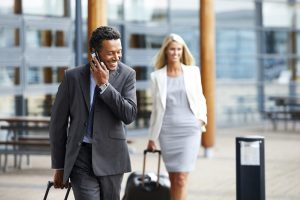 Business man walking with luggage outdoors using cell phone with woman in background